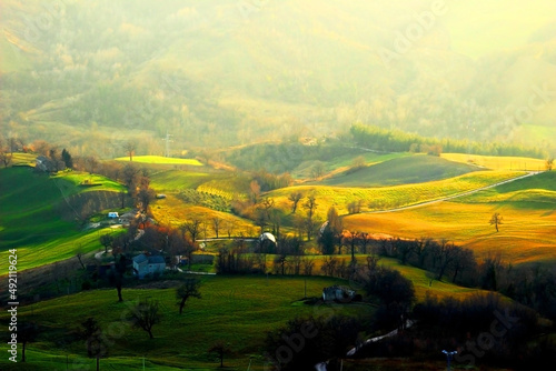 View from top on a hilly landscape in Penna San Giovanni with green fields partly illuminated by sunshine in the foreground and dense forests covered with fog in the background