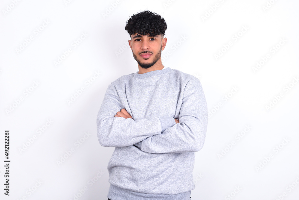 Serious pensive young arab man with curly hair wearing sport sweatshirt over white background feel like cool confident entrepreneur cross hands. Serious pensive young arab man with curly hair wearing sport sweatshirt over white background feel like cool confident entrepreneur cross hands.
