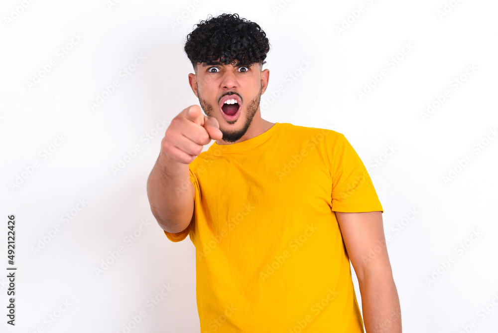 young arab man wearing yellow T-shirt over white background pointing displeased and frustrated to the camera, angry and furious ready to fight with you. young arab man wearing yellow T-shirt over white background pointing displeased and frustrated to the camera, angry and furious ready to fight with you.