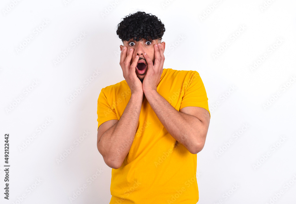 Stupefied young arab man wearing yellow T-shirt over white background expresses excitement and thrill, keeps jaw dropped, hands on cheeks, has eyes popped out Stupefied young arab man wearing yellow T-shirt over white background expresses excitement and thrill, keeps jaw dropped, hands on cheeks, has eyes popped out