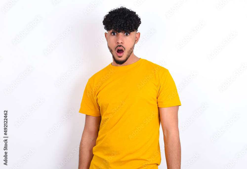 Fototapeta premium Oh my God. Surprised young arab man wearing yellow T-shirt over white background stares at camera with shocked expression exclaims with unexpectedness,