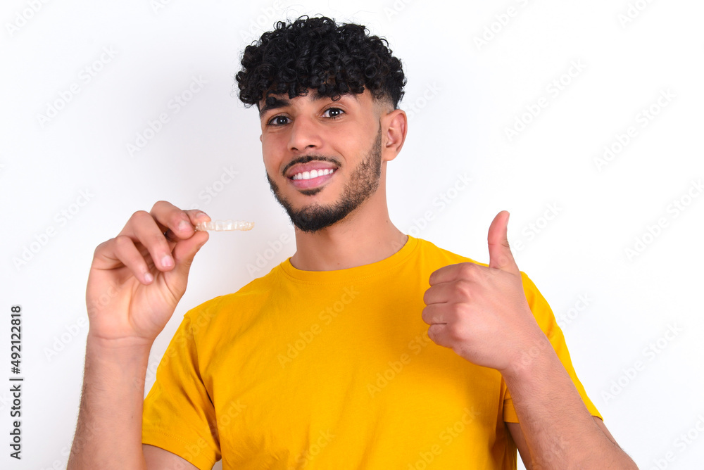 young arab man wearing yellow T-shirt over white background holding an invisible braces aligner and rising thumb up, recommending this new treatment. Dental healthcare concept. young arab man wearing yellow T-shirt over white background holding an invisible braces aligner and rising thumb up, recommending this new treatment. Dental healthcare concept.