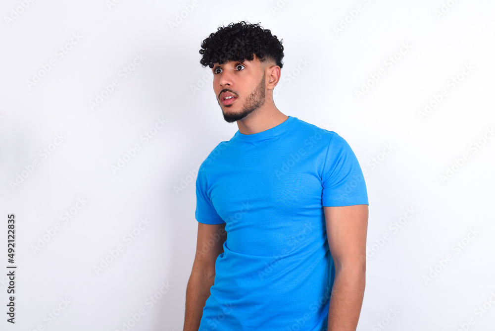 young arab man with curly hair wearing blue t-shirt over white background stares aside with wondered expression has speechless expression. Embarrassed model looks in surprise young arab man with curly hair wearing blue t-shirt over white background stares aside with wondered expression has speechless expression. Embarrassed model looks in surprise