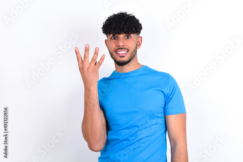 young arab man with curly hair wearing blue t-shirt over white background smiling and looking friendly, showing number four or fourth with hand forward, counting down