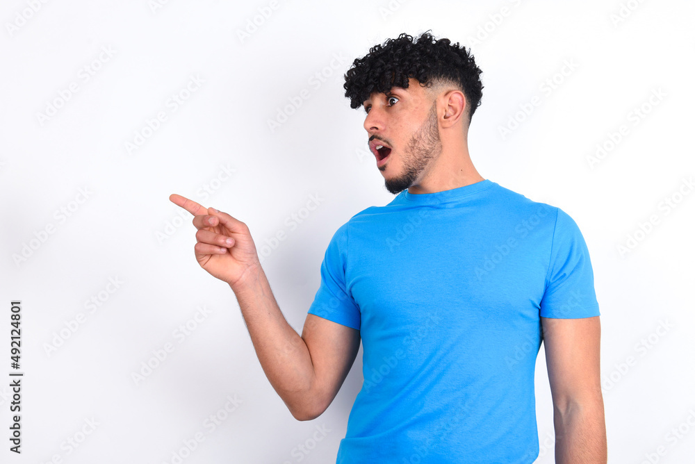 Stunned young arab man with curly hair wearing blue t-shirt over white background with greatly surprised expression points away on copy space, indicates something Stunned young arab man with curly hair wearing blue t-shirt over white background with greatly surprised expression points away on copy space, indicates something