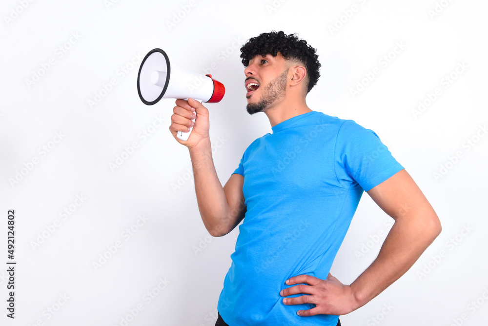 Funny young arab man with curly hair wearing blue t-shirt over white background People sincere emotions lifestyle concept. Mock up copy space. Screaming in megaphone. Funny young arab man with curly hair wearing blue t-shirt over white background People sincere emotions lifestyle concept. Mock up copy space. Screaming in megaphone.