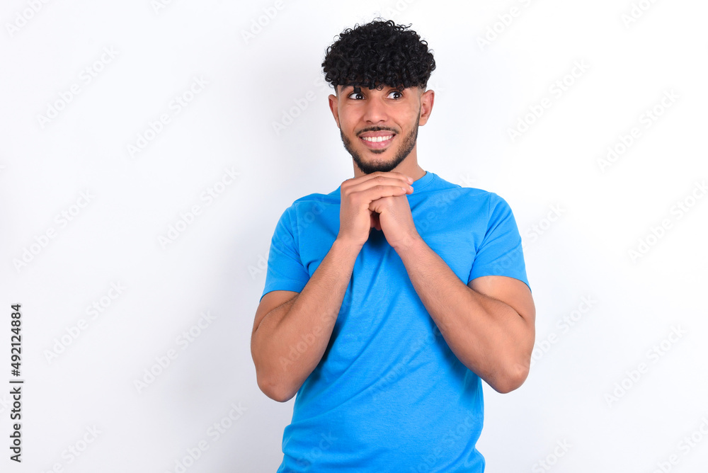 Happy young arab man with curly hair wearing blue t-shirt over white background anticipates something awesome happen, looks happily aside, keeps hands together near face, has glad expression. Happy young arab man with curly hair wearing blue t-shirt over white background anticipates something awesome happen, looks happily aside, keeps hands together near face, has glad expression.
