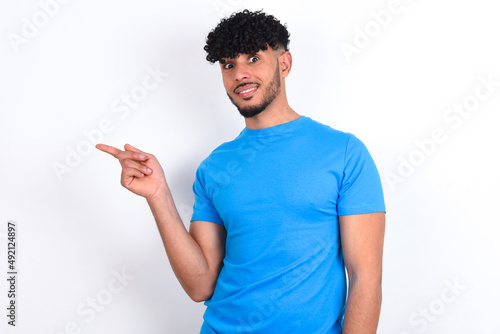 young arab man with curly hair wearing blue t-shirt over white background points to side on blank space demonstrates advertisement. People and promotion concept