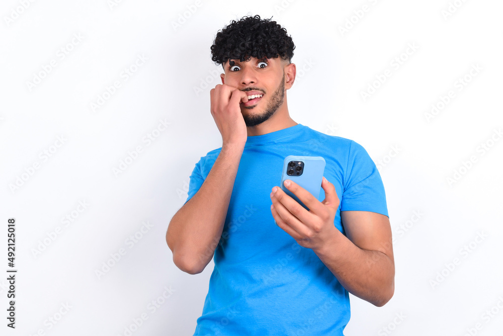 Afraid funny young arab man with curly hair wearing blue t-shirt over white background holding telephone and bitting nails Afraid funny young arab man with curly hair wearing blue t-shirt over white background holding telephone and bitting nails