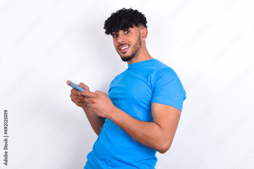 Nice addicted cheerful young arab man with curly hair wearing blue t-shirt over white background using gadget playing network game Nice addicted cheerful young arab man with curly hair wearing blue t-shirt over white background using gadget playing network game