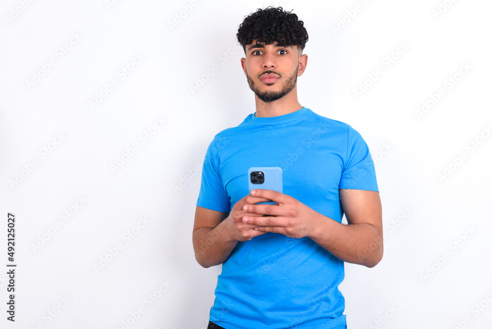 Portrait of serious confident young arab man with curly hair wearing blue t-shirt over white background holding phone in two hands Portrait of serious confident young arab man with curly hair wearing blue t-shirt over white background holding phone in two hands