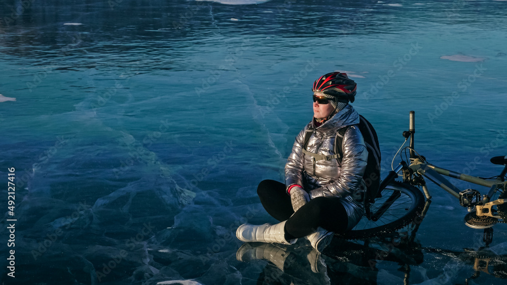 Woman sitting near her bike on ice. The girl cyclist stopped to rest. She sits on the wheel and enjoys the beautiful view of the sunset. The traveler is ride a cycle. Female is riding bike on the ice. Woman sitting near her bike on ice. The girl cyclist stopped to rest. She sits on the wheel and enjoys the beautiful view of the sunset. The traveler is ride a cycle. Female is riding bike on the ice.