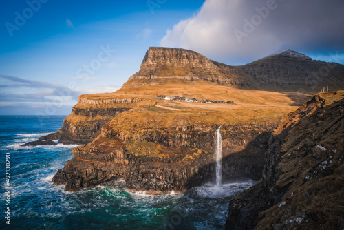 Sunny view of Mulafossur waterfall with Gasadalur village on background. Vagar island, Faroe Islands, Denmark. November 2021. Lonf exposure picture