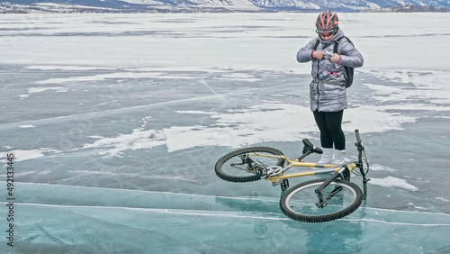 Woman stands near her bike on ice. The girl cyclist stopped to rest. The beautiful view of the sunset. The traveler is ride a cycle. Female is riding bike on the ice. Athlete puts on gear.