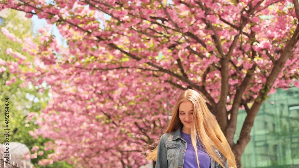 Beautiful young ukrainian girl feels free and happy look around and smiling on blurred background in the city street. Sakura tree. Caucasian woman with long hair walking around Cherry blossom trees. Beautiful young ukrainian girl feels free and happy look around and smiling on blurred background in the city street. Sakura tree. Caucasian woman with long hair walking around Cherry blossom trees.