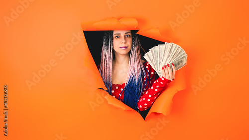 Bright woman with money sticks out of hole of orange background. Impressive female with dollar bills. © Anton Dios