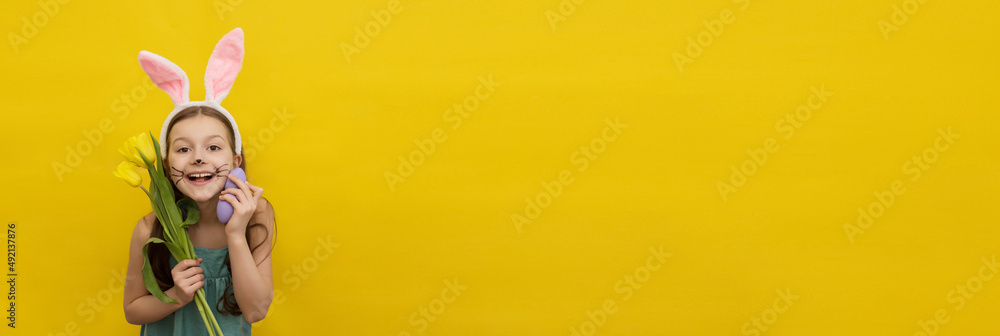 Little happy girl with long dark hair in a headband with rabbit ears holds colorful purple Easter eggs and yellow tulips near her face and looks at the camera on a yellow background, easter card