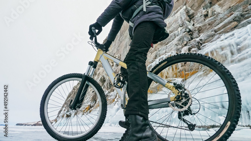 Man is riding bicycle near ice grotto. Rock with ice caves icicles. Teenage is dressed in black down jacket  cycling backpack  helmet. Tires on covered with special spikes. Traveler boy is ride cycle.