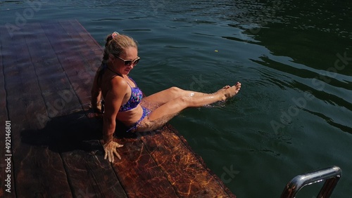 Woman sit on a pier in sunglasses and swimming suit. Girl rest on a flood wood underwater pier. The pavement is covered with water in lake. Lady in the water and splashes feet. View from above.