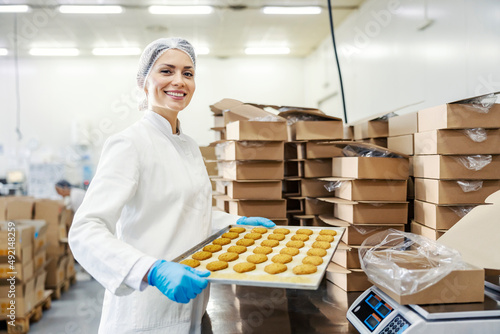 A female food factory worker holding tray with fresh cookies and smiling at the camera.