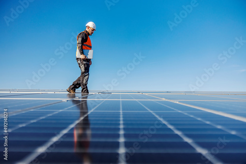 A worker walking around roof with solar panels with laptop under armpit.