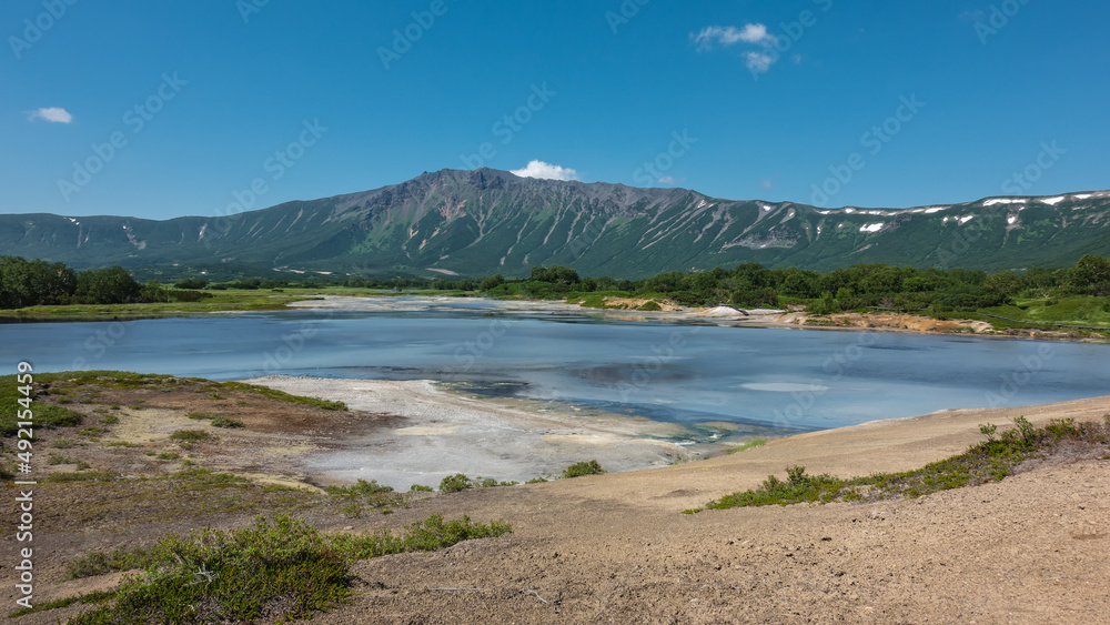 Turquoise thermal lake in the caldera of an extinct volcano. There are sulfur deposits on the soil. Green vegetation on the shore. A picturesque mountain range against the blue sky. Kamchatka. Uzon