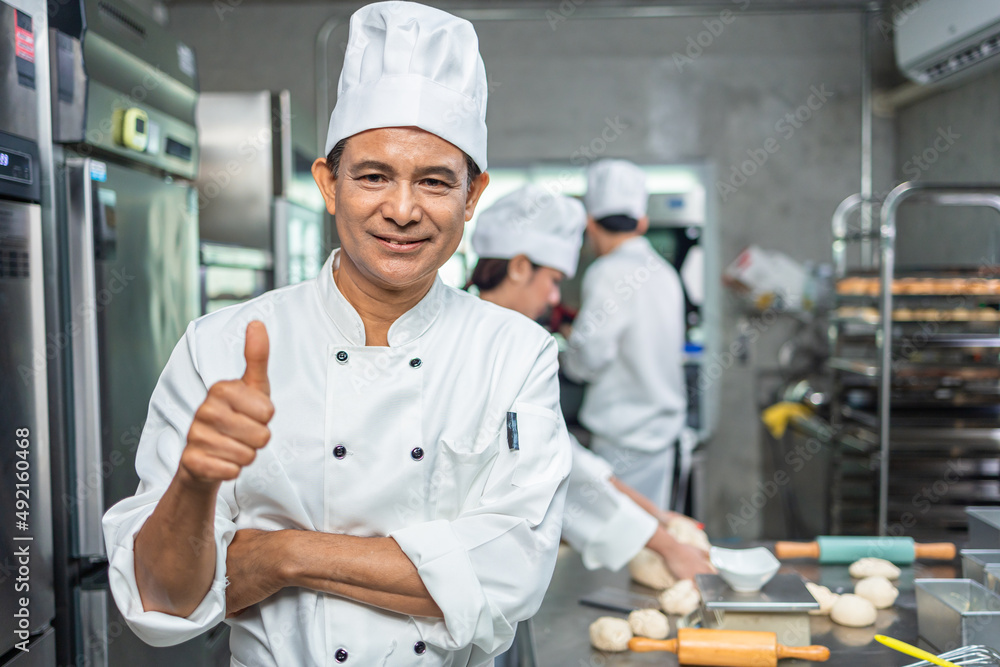 A senior Asian chef male  bakers in a white chef dresses uniform and hats standing cutting on a counter with many baked and unbaked bread on trays at a bakery kitchen restaurant.