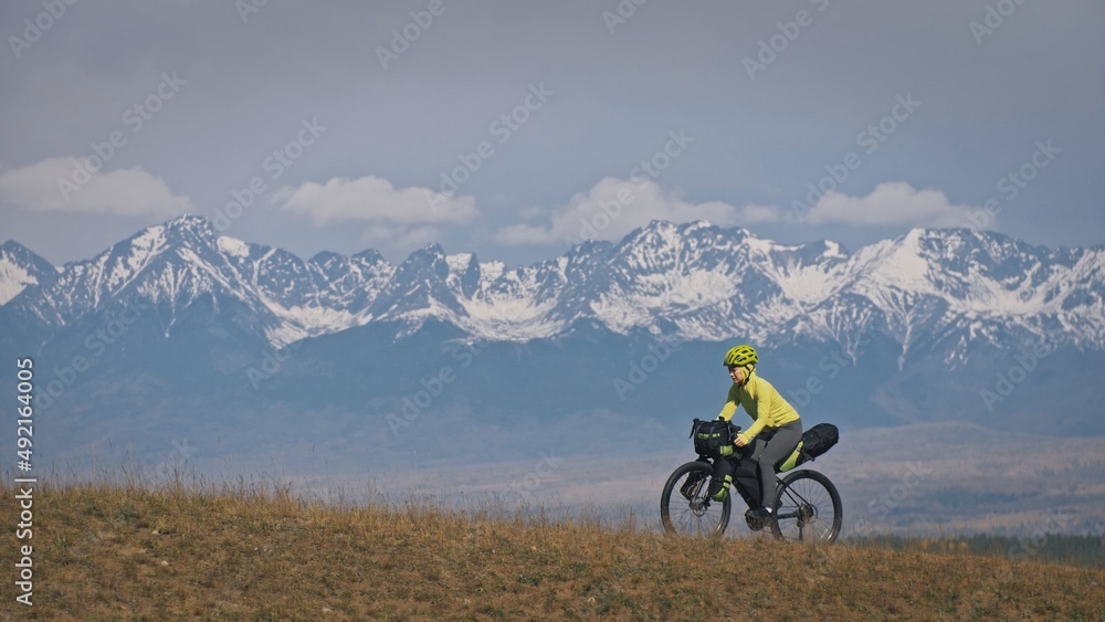 The woman travel on mixed terrain cycle touring with bikepacking. The traveler journey with bicycle bags. Sport tourism bikepacking, bike, sportswear in green black colors. Mountain snow capped. The woman travel on mixed terrain cycle touring with bikepacking. The traveler journey with bicycle bags. Sport tourism bikepacking, bike, sportswear in green black colors. Mountain snow capped.