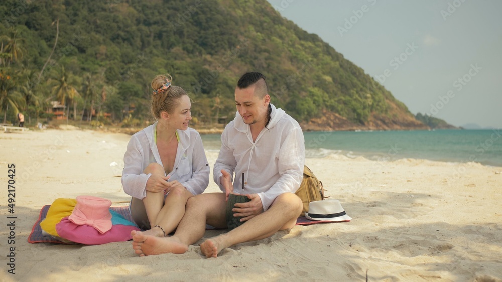 The cheerful love couple holding and eating slices of watermelon on tropical sand beach sea. Romantic lovers two people caucasian spend summer weekend in outdoor. Hat, backpack white shirt beachwear.