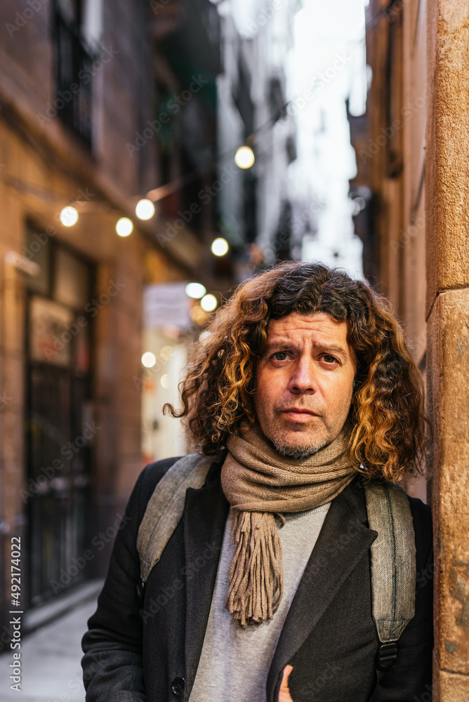 Serious middle aged male in smart casual clothes with backpack and scarf looking at camera while leaning on wall at dusk on city street Serious middle aged male in smart casual clothes with backpack and scarf looking at camera while leaning on wall at dusk on city street