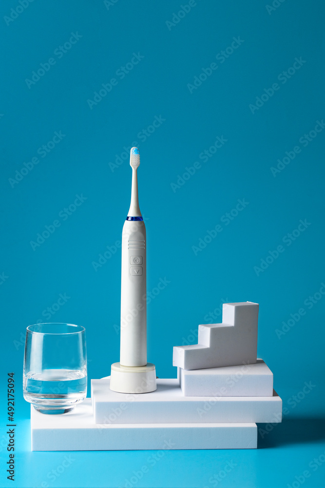 Stomatology clinic and dental care still life concept.Electric Toothbrush and glass on water on podiums on blue background Stomatology clinic and dental care still life concept.Electric Toothbrush and glass on water on podiums on blue background