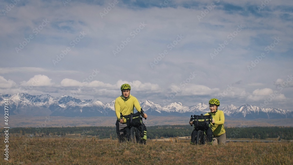 Obraz premium The man and woman travel on mixed terrain cycle touring with bikepacking. The two people journey with bicycle bags. Sport bikepacking, bike, sportswear in green black colors. Mountain snow capped.