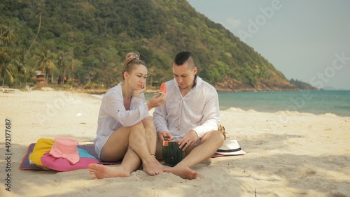 The cheerful love couple holding and eating slices of watermelon on tropical sand beach sea. Romantic lovers two people caucasian spend summer weekend in outdoor. Hat  backpack white shirt beachwear.