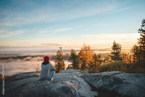 Hiker sits atop Vuokatinvaara in Sotkamo in the Kainuu region, Finland at sunrise and above the clouds. A brunette woman with a red knitted hat on top of the mountain
