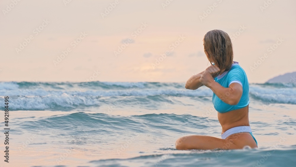 Fototapeta premium SLOW MOTION GLOW MIST GOLD SUNSET CINEMATIC VIEW: Attractive woman sitting on water sea. Girl in turquoise swimsuit. Freedom paradise holiday vacation summer beach and seaside landscape concept