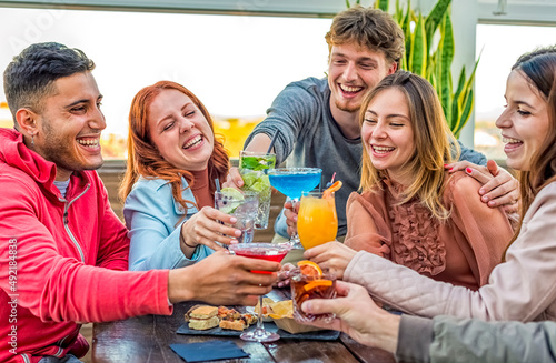 young adult group of friends having cocktail on a rooftop bar restaurant outdoors. people enjoying happy hour together drinking alcohol and eating sitting on a table. fun and lifestyle concept