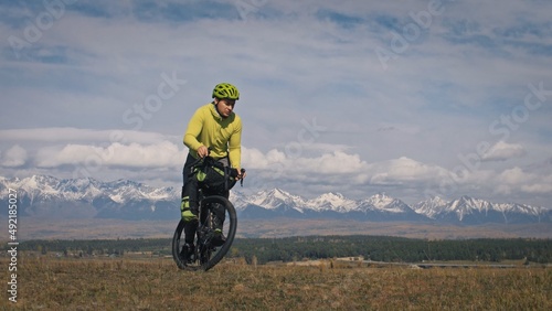 Fototapeta Naklejka Na Ścianę i Meble -  The man travel on mixed terrain cycle touring with bikepacking. The traveler journey with bicycle bags. Sport tourism bikepacking, bike, sportswear in green black colors. Mountain snow capped.