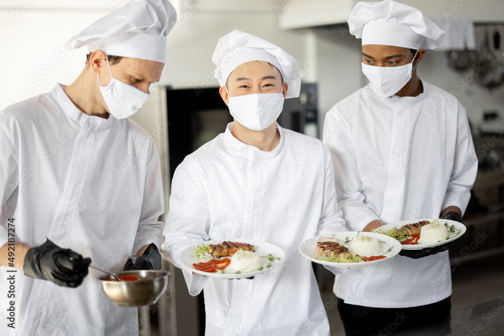 Portrait of multiracial team of cooks standing with ready meals for a restaurant in the kitchen. Chefs wearing uniform and face mask during pandemic. Asian chef with Latin and European guys