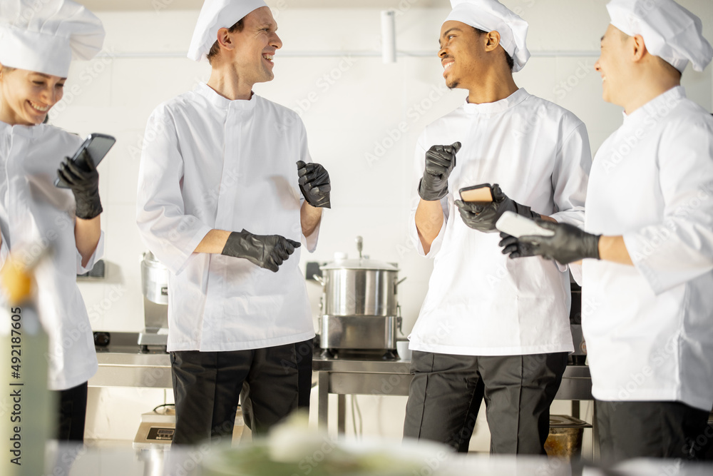 Team of multiracial cooks having conversation during a coffee break in the kitchen. Well-dressed chefs resting and having fun in the restaurant kitchen. Teamwork and leisure time on work Team of multiracial cooks having conversation during a coffee break in the kitchen. Well-dressed chefs resting and having fun in the restaurant kitchen. Teamwork and leisure time on work