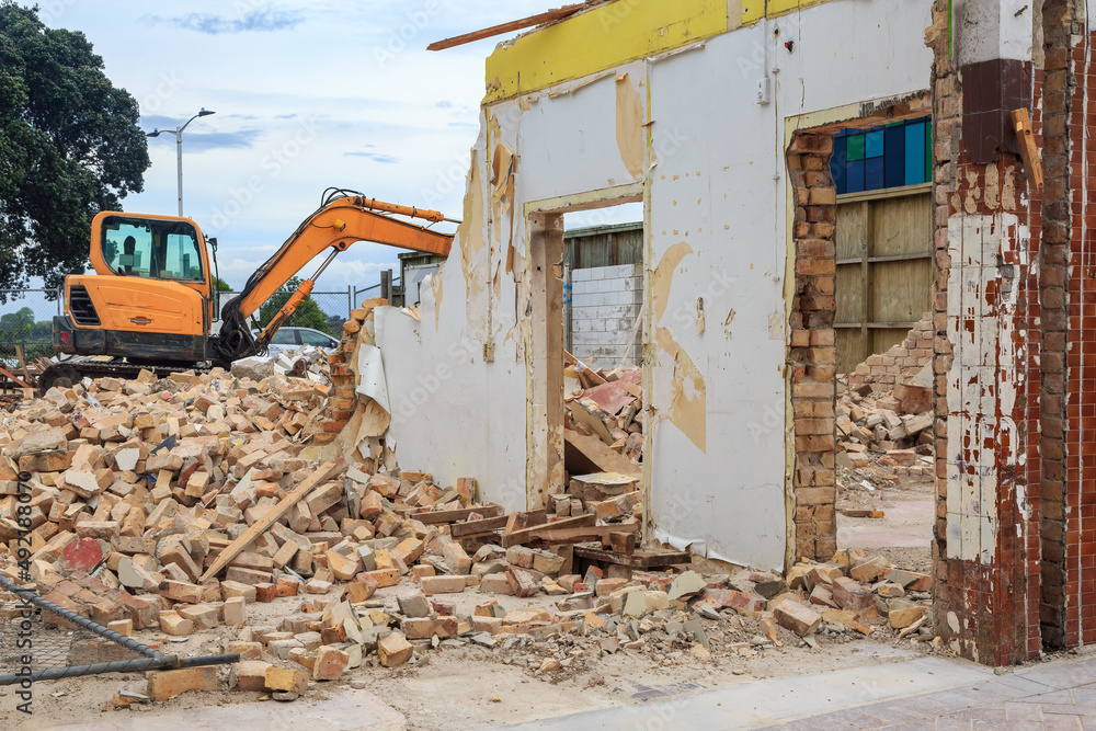 A building being demolished, with an excavator in the background A building being demolished, with an excavator in the background