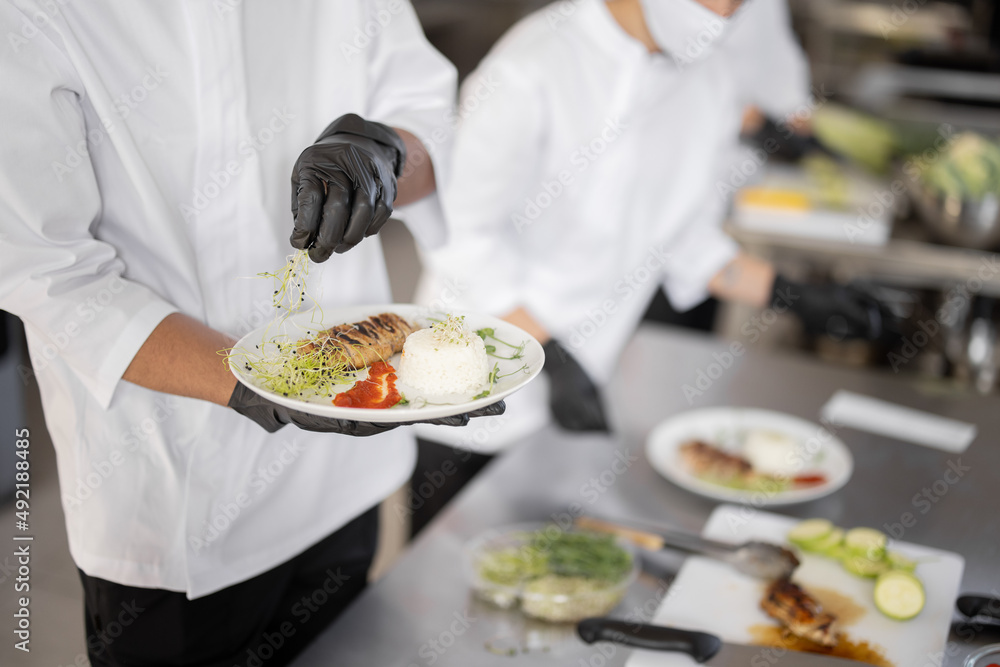 Cooks finishing main courses, putting greens on plates with rise and meat, close-up on table with meals in the kitchen. Chefs wearing gloves and uniform. Concept of haute cuisine and professional