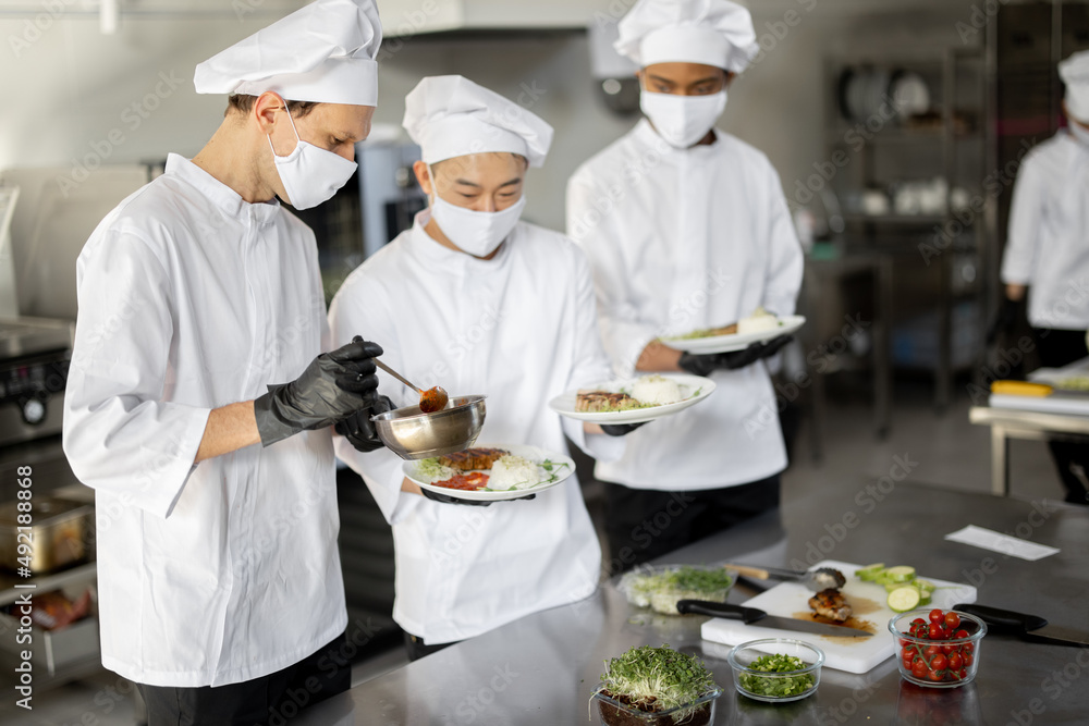 Multiracial team of cooks standing with ready meals for a restaurant in the kitchen. Chefs wearing uniform and face mask during pandemic. Asian chef with Latin and European guys Multiracial team of cooks standing with ready meals for a restaurant in the kitchen. Chefs wearing uniform and face mask during pandemic. Asian chef with Latin and European guys