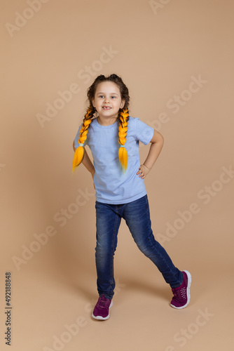 Excited ,small girl dancing by moving legs smiling looking away having yellow kanekalon braids on head on beige background wearing blue t-shirt and jeans.