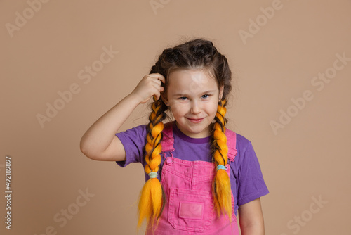 Portrait of young, smiling girl with kanekalon braids of yellow color touching hair with hand looking at camera wearing pink jumpsuit and purple t-shirt on beige background.