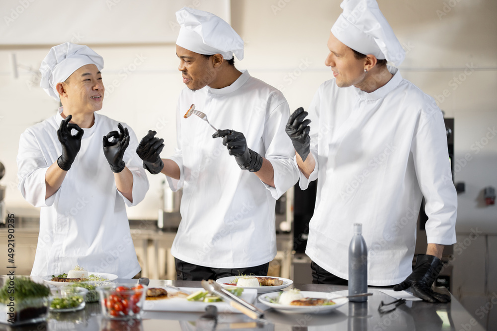 Three cooks with different nationalties tasting cooked meals, inventing new dishes for a menu. Caucasian chef with Latin and Asian cooks in uniform working together. Concept of teamwork and high