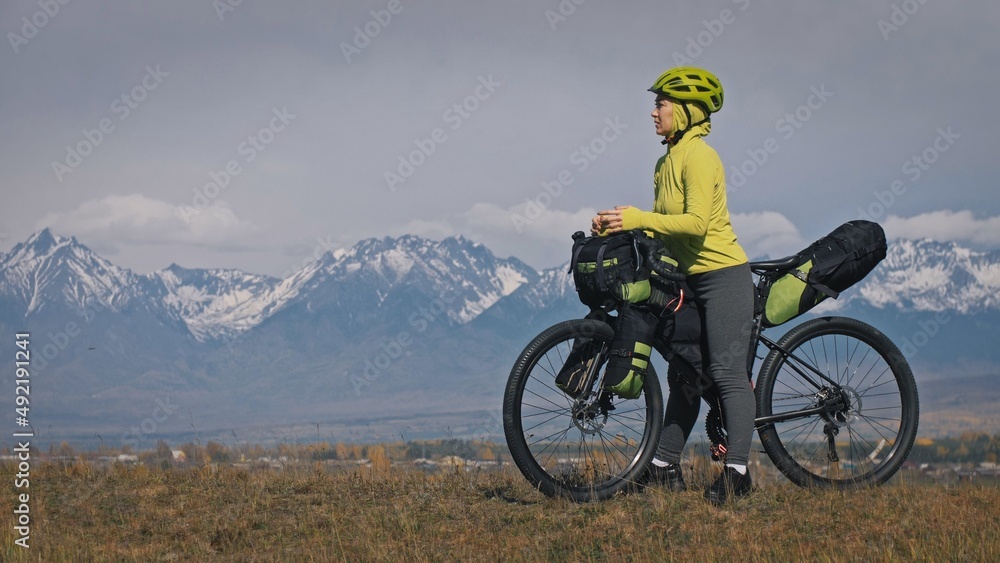 The woman travel on mixed terrain cycle touring with bikepacking. The traveler journey with bicycle bags. Sport tourism bikepacking, bike, sportswear in green black colors. Mountain snow capped. The woman travel on mixed terrain cycle touring with bikepacking. The traveler journey with bicycle bags. Sport tourism bikepacking, bike, sportswear in green black colors. Mountain snow capped.