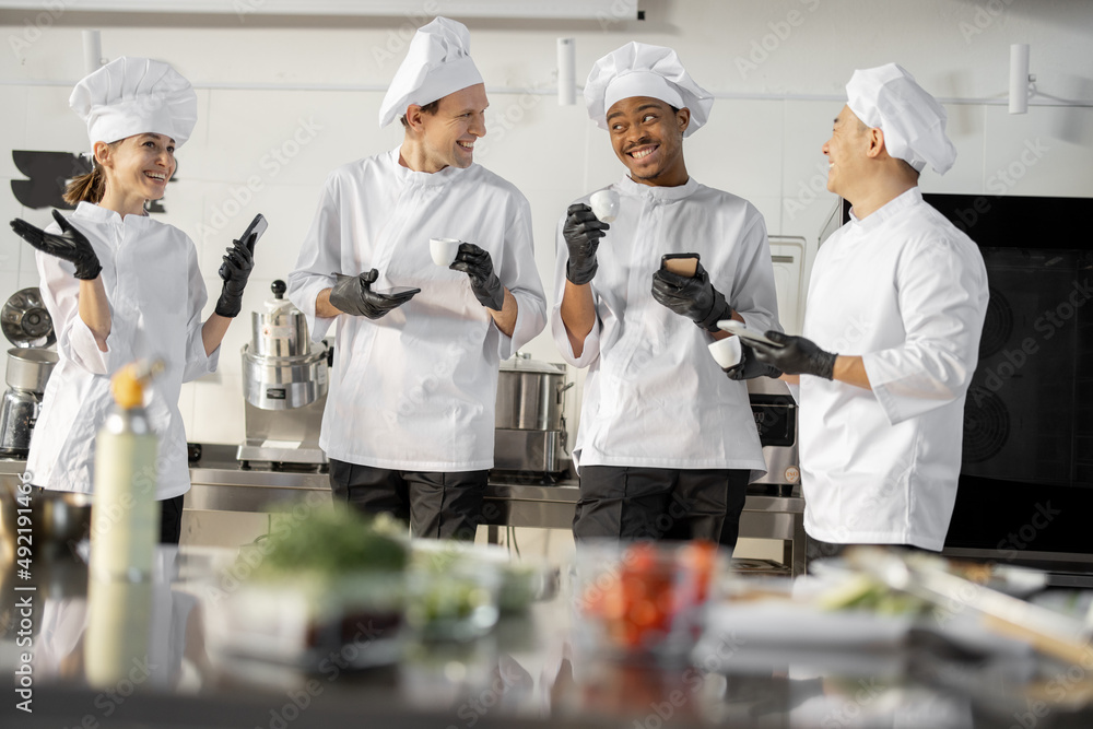 Team of multiracial cooks having conversation during a coffee break in the kitchen. Well-dressed chefs resting and having fun in the restaurant kitchen. Teamwork and leisure time on work
