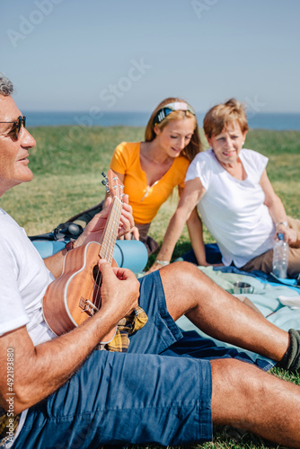 Senior man playing ukulele for his family sitting on a blanket during an excursion outdoors. Selective focus on man in foreground © David Pereiras