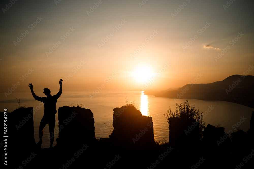 Fototapeta premium A woman or a man standing on a rock and looking directly at the sunset by the sea. The concept of nature and beauty. Orange sunset. Silhouette at sunset