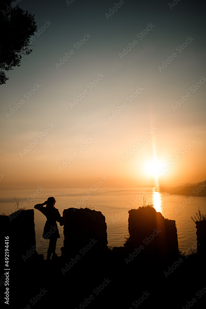 Fototapeta premium A woman or a man standing on a rock and looking directly at the sunset by the sea. The concept of nature and beauty. Orange sunset. Silhouette at sunset